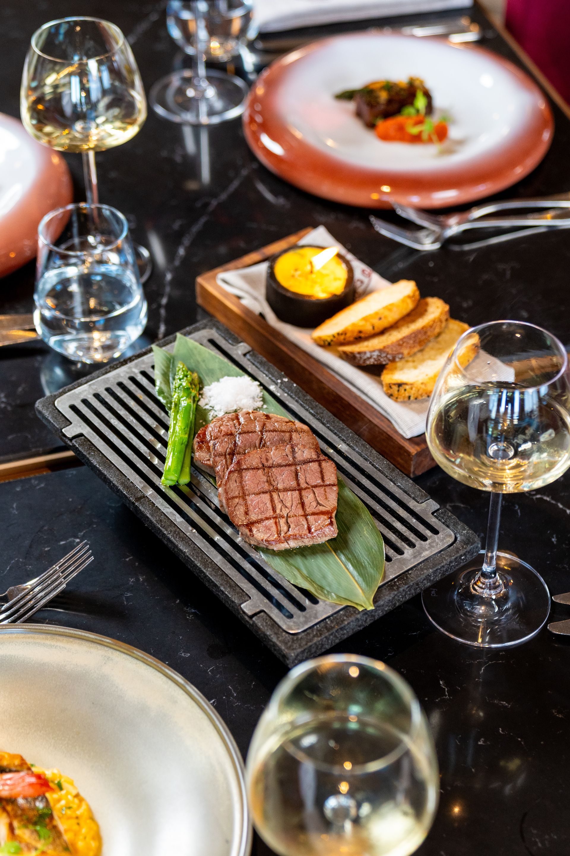Liu Resorts | Steak dinner with asparagus, bread, and wine glasses on a black marble table.