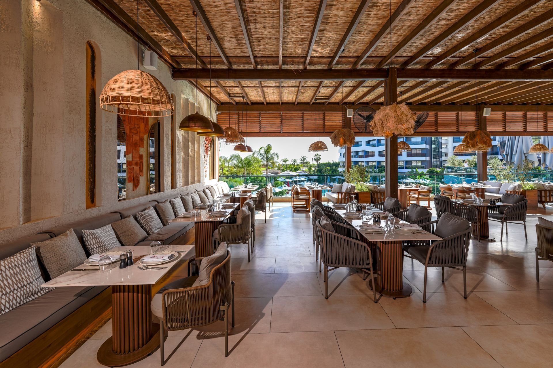 Liu Resorts | Restaurant interior with tables, woven chairs, and a long built-in bench. Wooden ceiling and outdoor view.