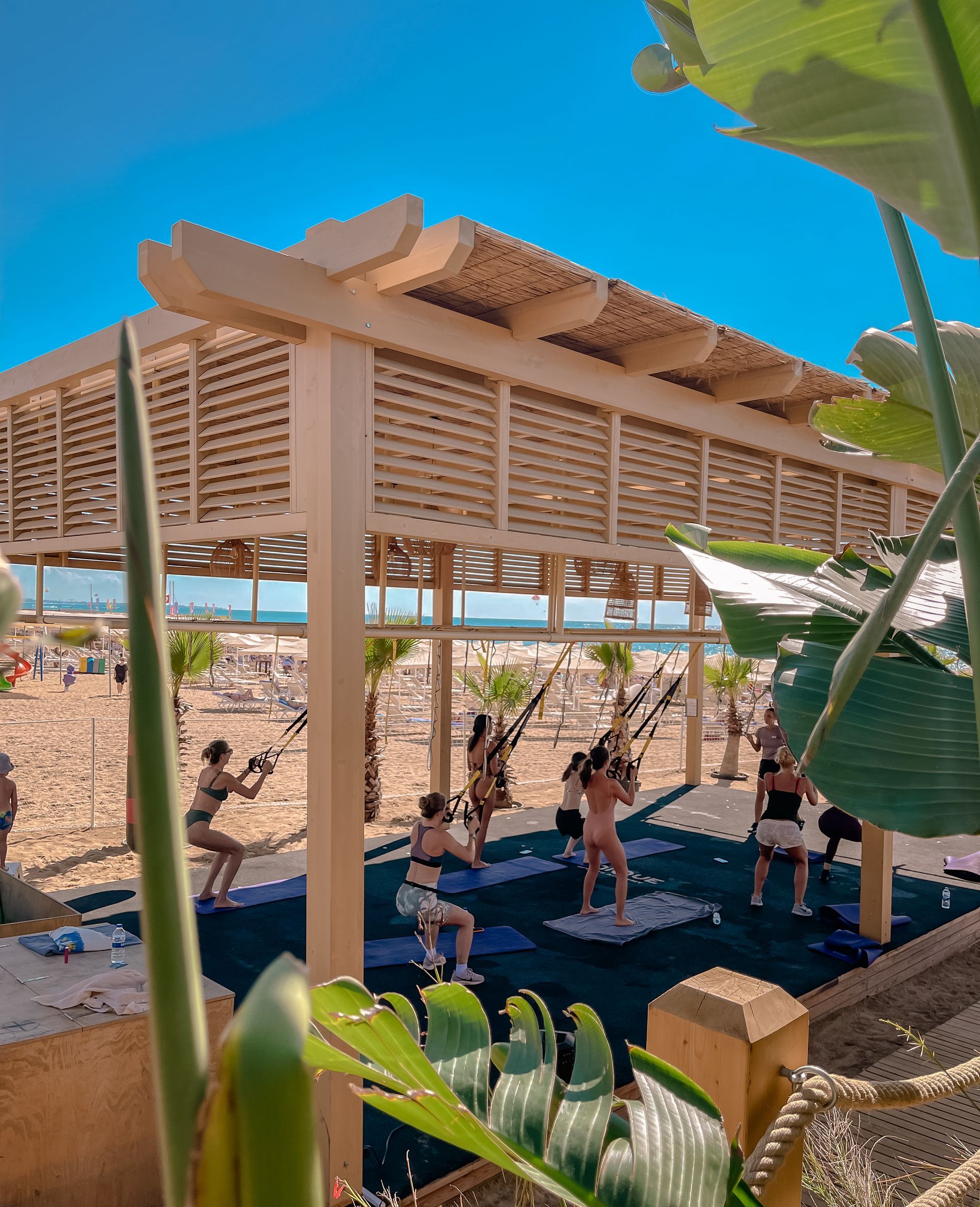 Liu Resorts | People exercising on a beach under a wooden structure, blue sky, tropical foliage.