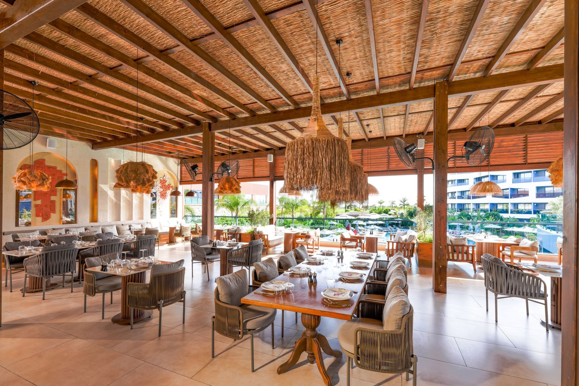 Restaurant with wooden structure, tables, chairs, and woven ceiling. View overlooks a pool area.