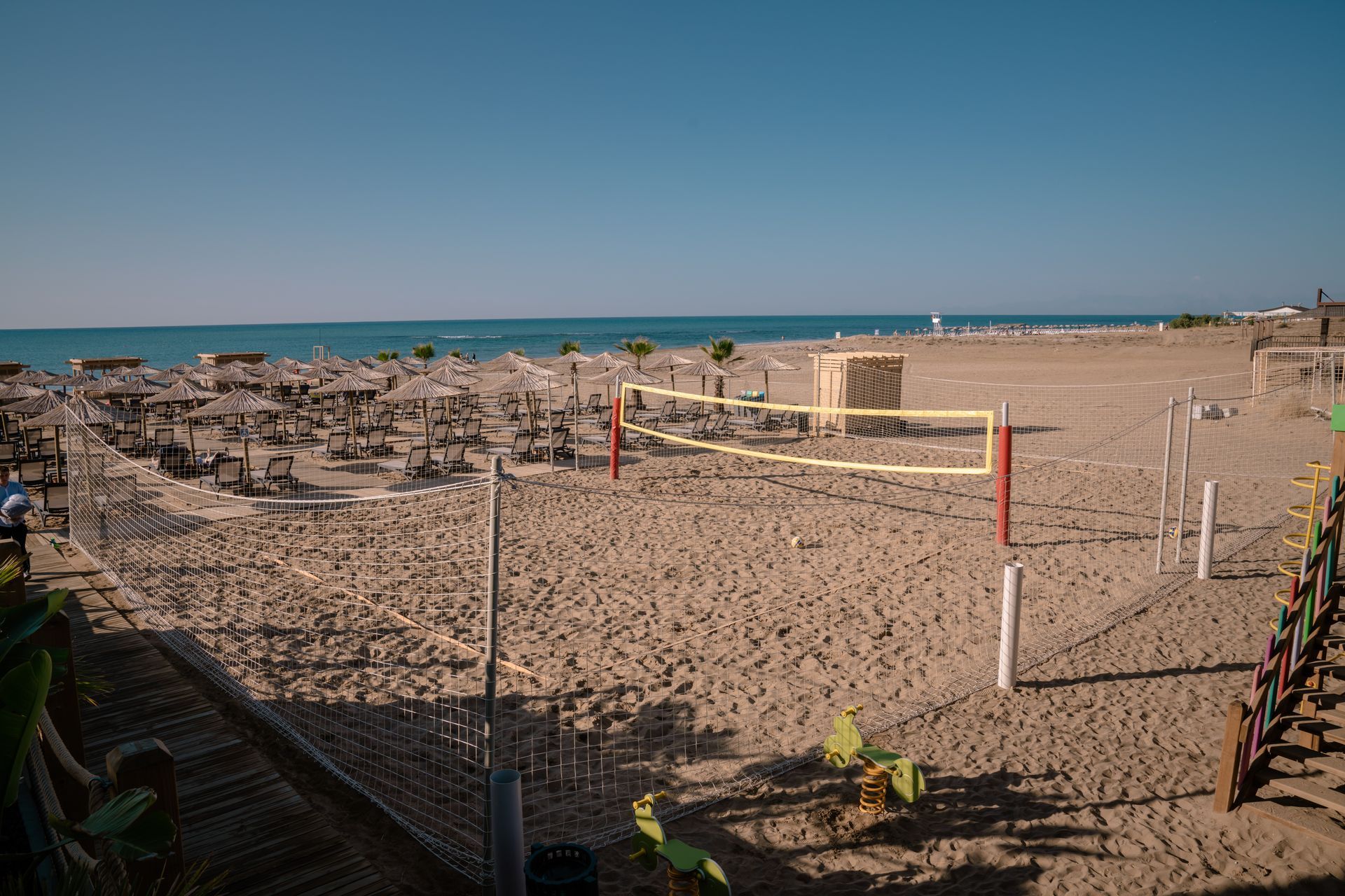 Liu Resorts | Beach with volleyball net; umbrellas and chairs line the shore; sunny day, blue sky.