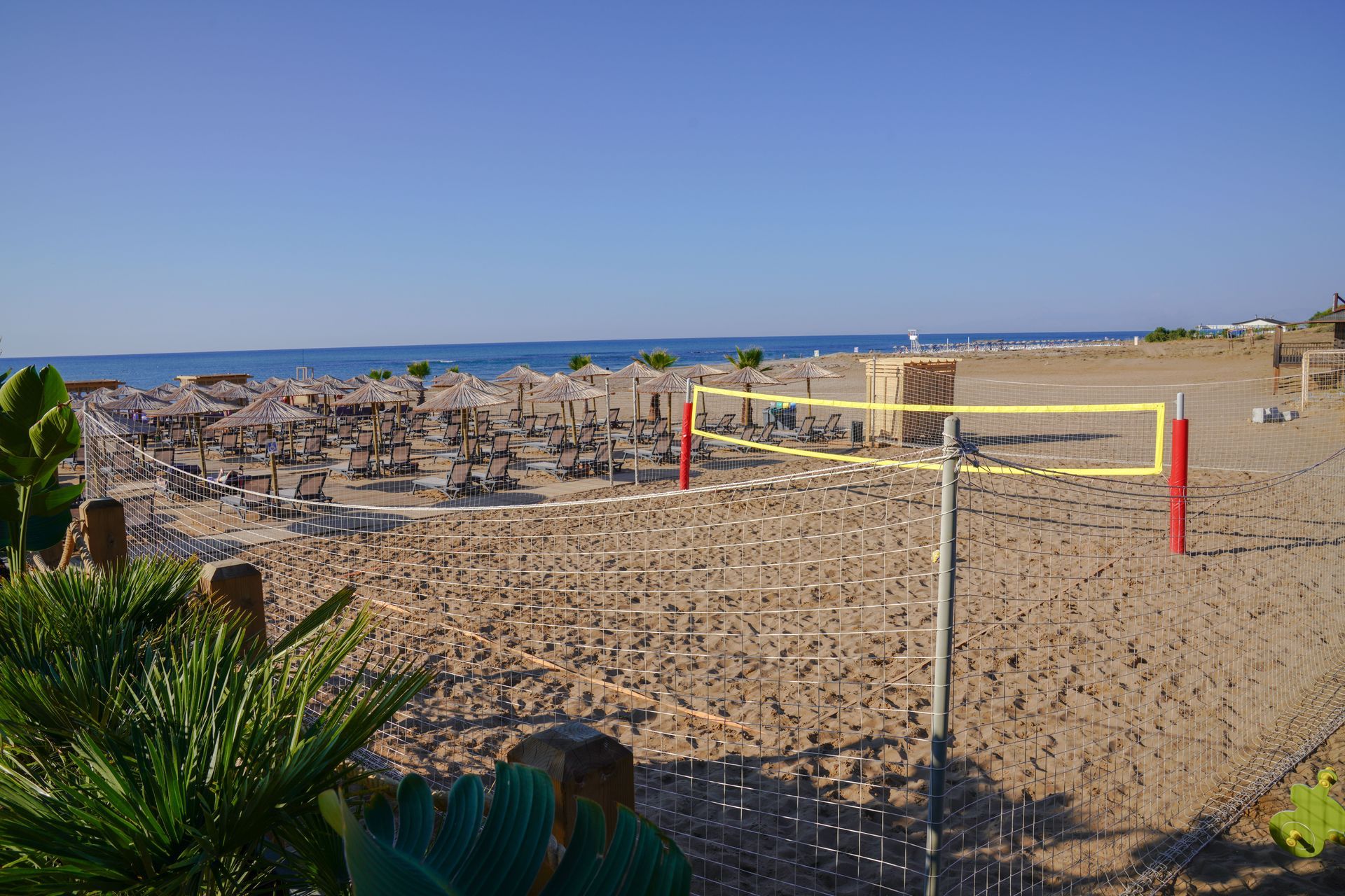 Strandszene mit Volleyballnetz im Vordergrund; Sonnenschirme und Strandbesucher im Hintergrund.