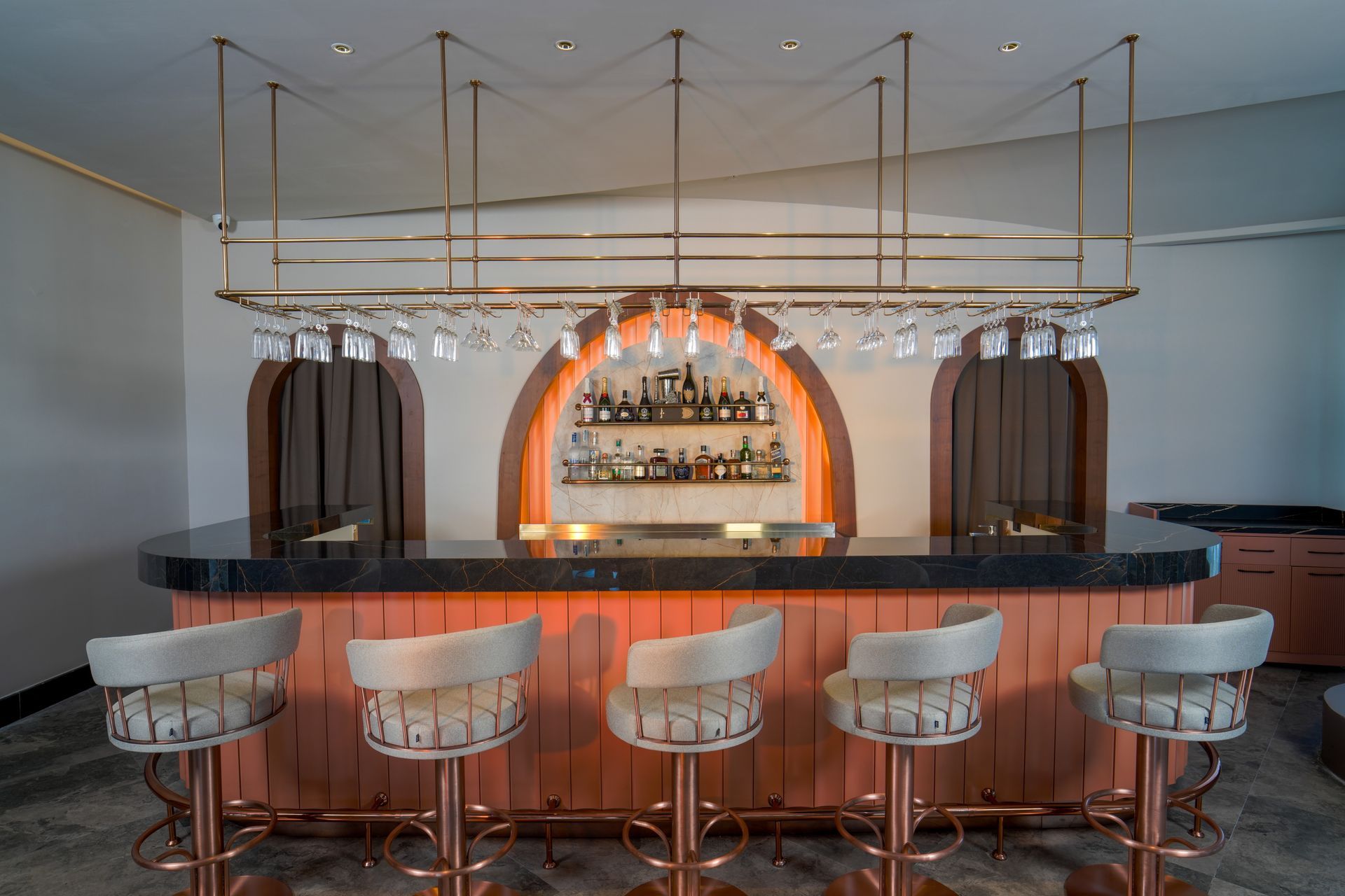 Bar with copper accents, curved counter, and five upholstered stools. Overhead rack holds wine glasses.