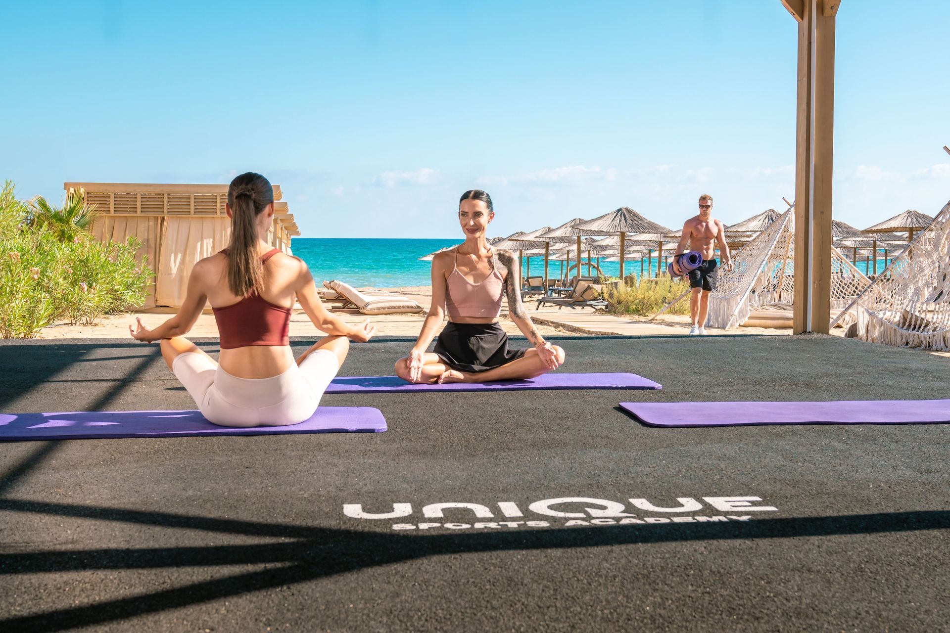 Liu Resorts | Two people in yoga poses on mats at beach, turquoise water visible. A man walks in background.
