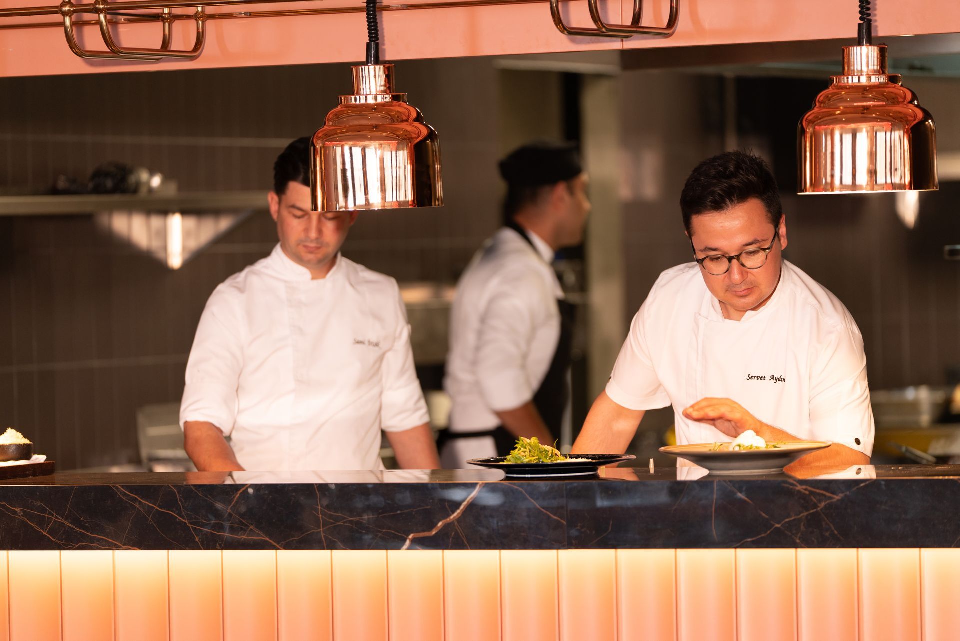 Liu Resorts | Chefs preparing food at a restaurant kitchen counter with copper pendant lights.