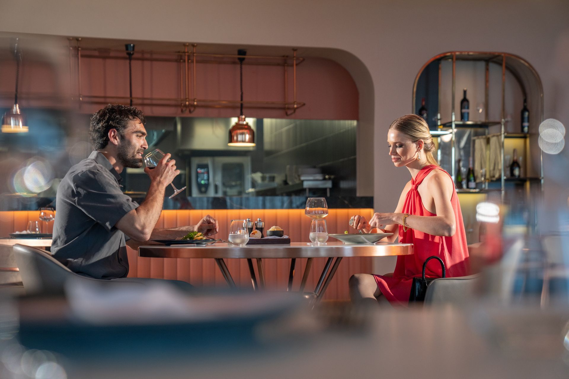 Liu Resorts | Couple at a restaurant table, man drinking wine, woman eating. Modern interior with pink accents.