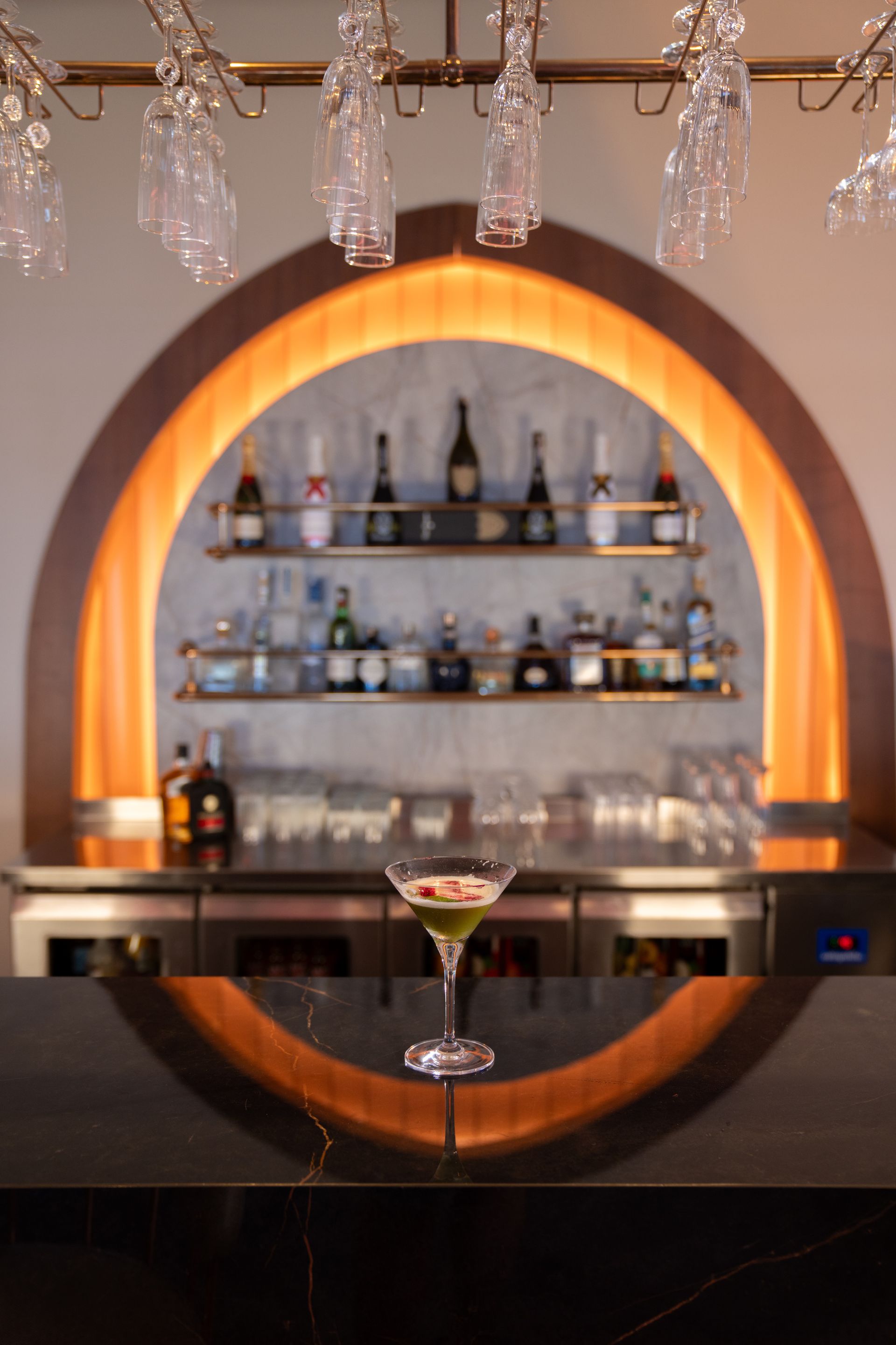 Liu Resorts | Cocktail on bar with arched shelving, bottles, and hanging glassware illuminated by warm lighting.