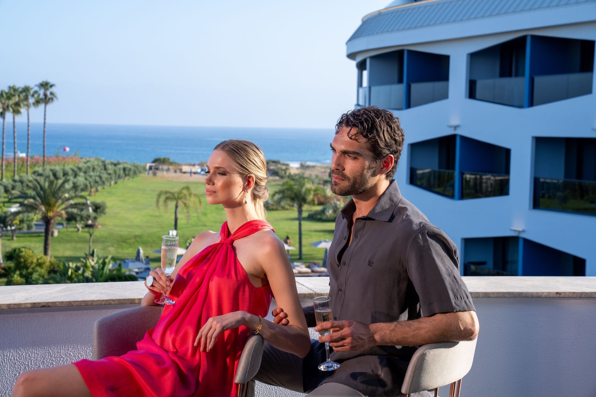 Liu Resorts | Woman in red dress and man with dark hair holding drinks, overlooking a coastal landscape.