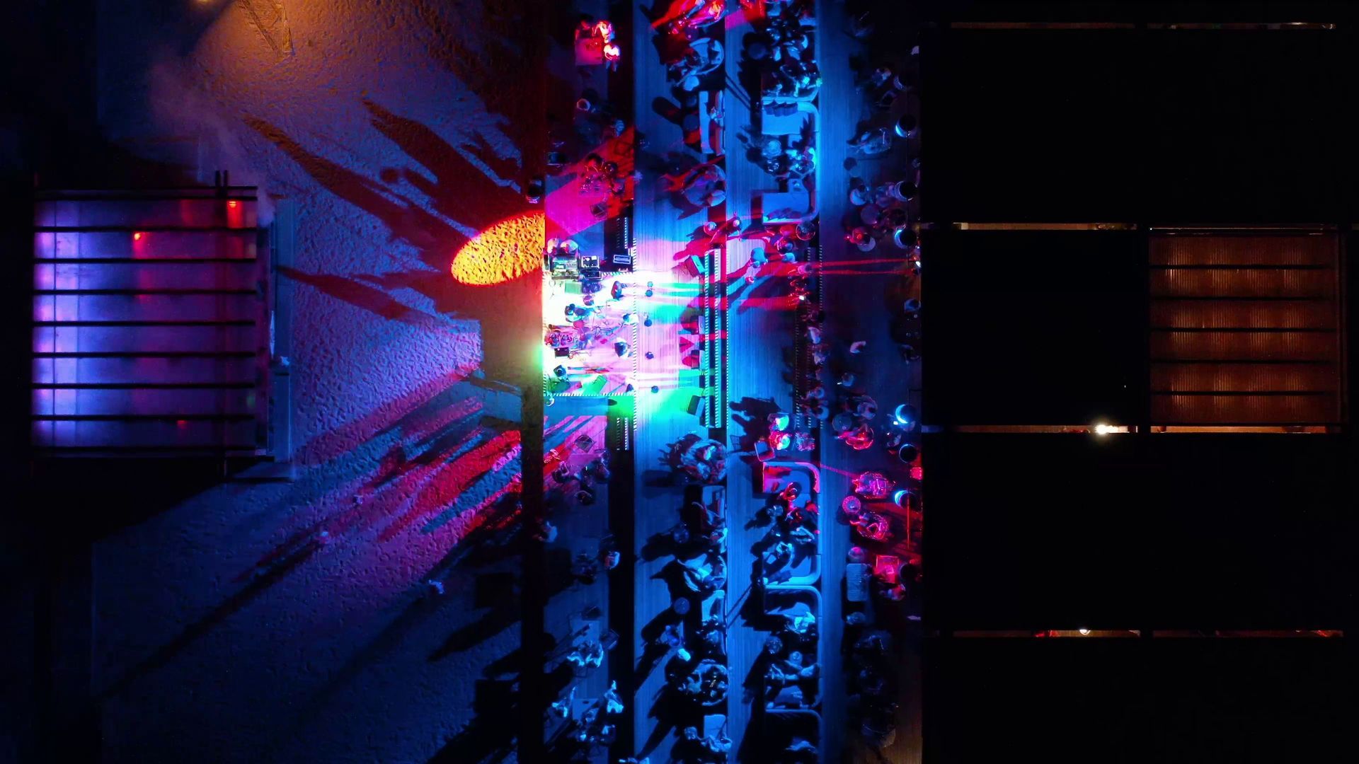 Overhead night view of a brightly lit street crowded with people, surrounded by buildings.